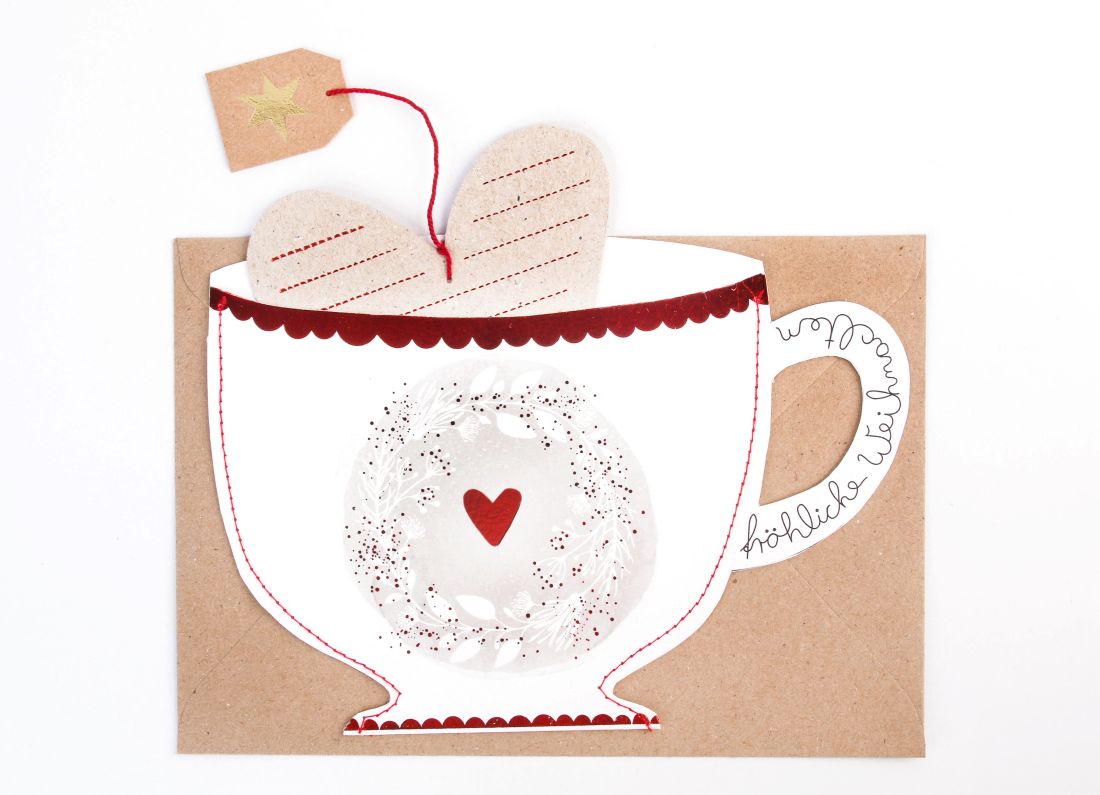 Teetassenförmige Weihnachtskarte in Weiß und Rot mit Blätterkranz, rotem Herz und dem Schriftzug Fröhliche Weihnachten am Tassenhenkel sowie herausnehmbarer Herzkarte an einer roten Schnur