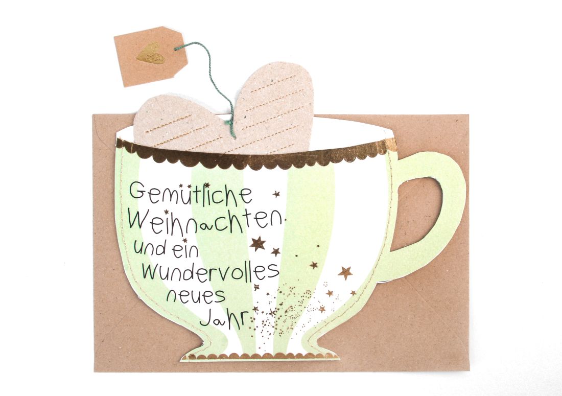 Teetassenförmige Weihnachtskarte in Hellgrün und Weiß mit dem Aufdruck Gemütliche Weihnachten und ein wundervolles neues Jahr sowie herausnehmbarer Herzkarte an einer Schnur